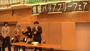写真１　講演会壇上で話す岩井さん。佐々木健、佐々木亜紀、手話通訳士が立っている。
